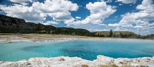 A vibrant blue hot spring surrounded by rocky terrain and lush greenery under a cloudy sky.