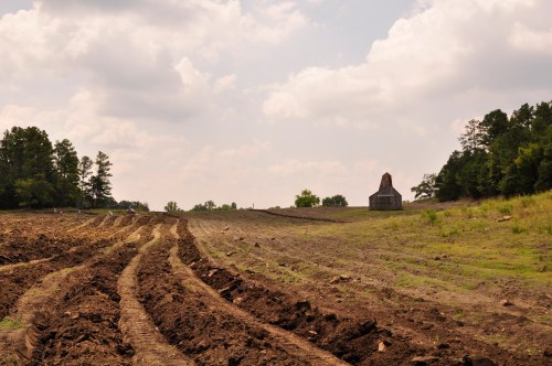 A plowed field stretches towards a distant barn under a cloudy sky, surrounded by trees.