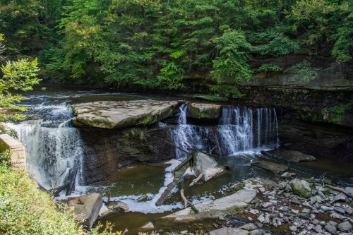 A serene waterfall cascades over rocky ledges, surrounded by lush green trees and a tranquil river.