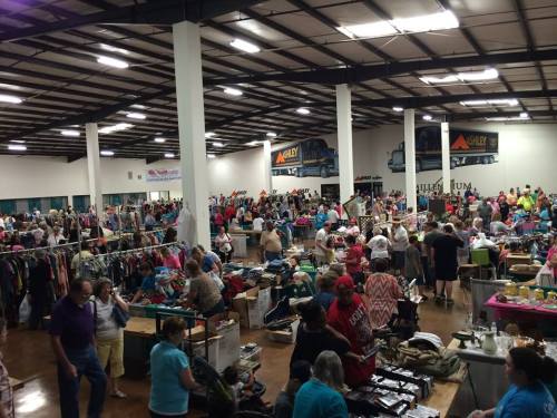 A bustling indoor market with many people browsing tables filled with clothes and various items. Brightly lit space.