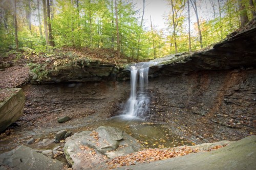 A serene waterfall cascades over rocky ledges, surrounded by lush green trees and autumn leaves on the ground.