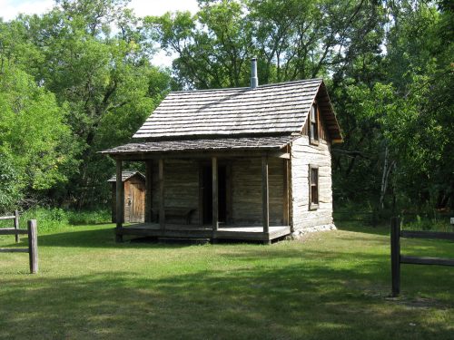 A rustic wooden cabin surrounded by trees, featuring a porch and a small outbuilding nearby.