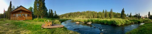 A panoramic view of a wooden cabin by a serene river, surrounded by lush greenery and tall trees under a clear blue sky.