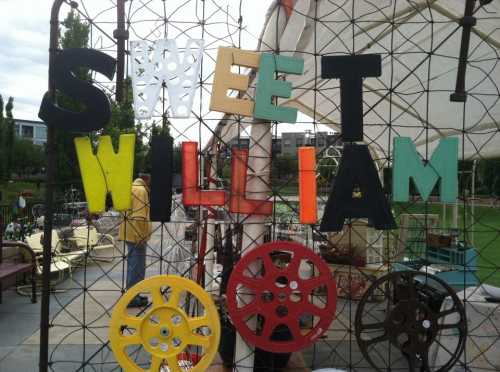 Colorful letters spelling "SWEET WILLIAM" on a wire fence, with vintage film reels below. Outdoor market setting.