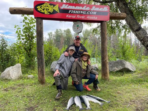 Three people pose with caught fish in front of a fishing sign at Kenai River, Alaska, surrounded by greenery.