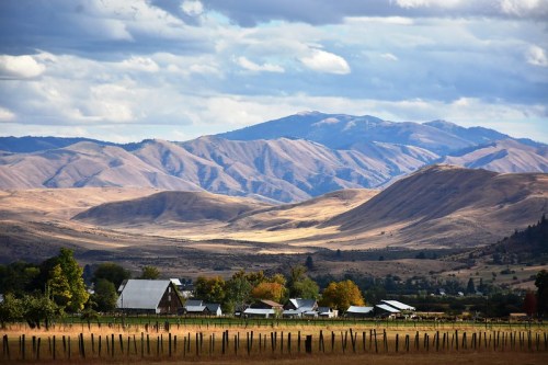 A scenic view of rolling hills and mountains under a cloudy sky, with a small farm in the foreground.