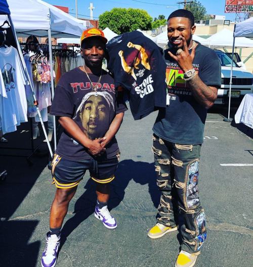 Two men pose outdoors, one holding a shirt featuring Tupac, surrounded by clothing stalls. Bright, sunny day.