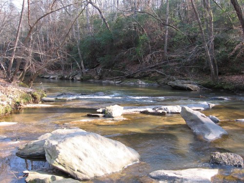 A serene river flows through a wooded area, with smooth rocks scattered along the water's edge.