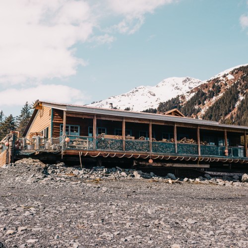 A wooden cabin on a rocky shore, surrounded by trees and mountains under a clear blue sky.