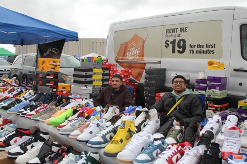 Two vendors sit behind a table filled with various sneakers and shoe boxes at an outdoor market.