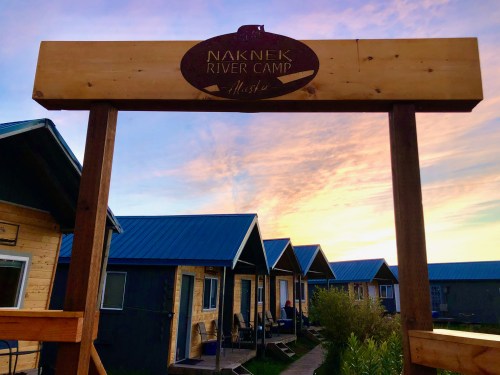 Sign for Naknek River Camp in Alaska, with cabins and a colorful sunset in the background.