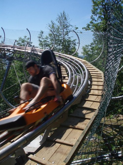 A person rides a roller coaster on a wooden track, surrounded by trees and a clear blue sky.