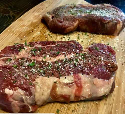 Two raw steaks seasoned with salt and herbs, resting on a wooden cutting board.