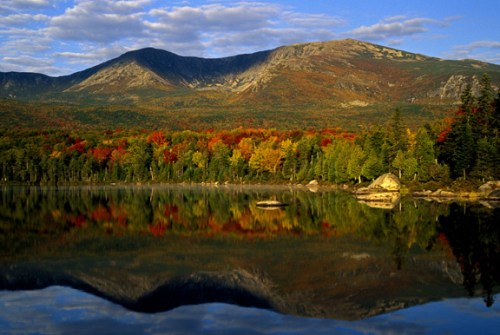 A serene lake reflects vibrant autumn foliage and mountains under a blue sky with scattered clouds.