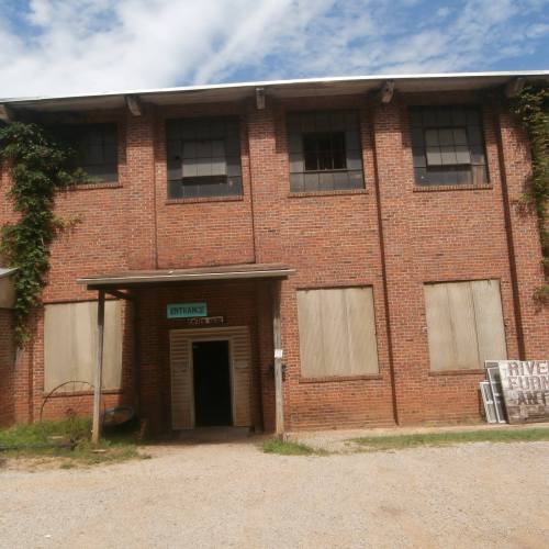 A brick building with boarded windows and a sign reading "Entrance" on the door, surrounded by greenery.