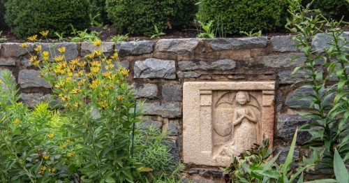 A stone relief of a figure in prayer set into a textured stone wall, surrounded by yellow flowers and greenery.