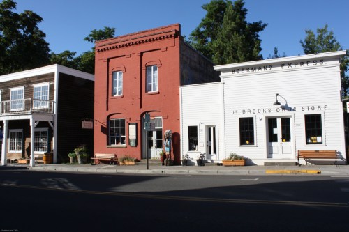 Historic buildings line a street, featuring a red brick structure and a white storefront with benches outside.