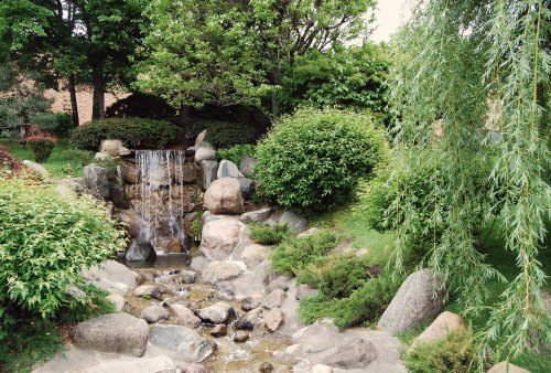 A serene garden scene featuring a small waterfall cascading over rocks, surrounded by lush greenery and trees.