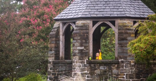 A child in a yellow raincoat waves from a stone gazebo surrounded by colorful trees and greenery.