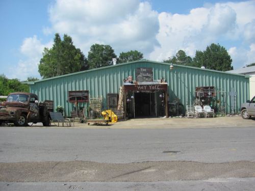 A green metal building with a sign reading "Hey Y'all," surrounded by vintage vehicles and various items outside.