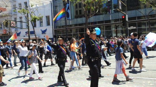 A vibrant parade scene with people celebrating, including police officers waving and rainbow flags flying.