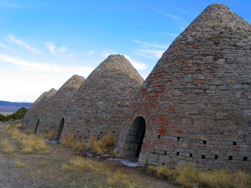 Four stone beehive-shaped kilns stand in a row against a blue sky, surrounded by dry grass and distant mountains.