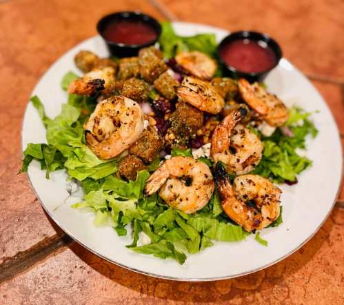 A plate of grilled shrimp on a bed of lettuce, accompanied by fried okra and two dipping sauces.