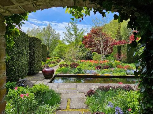 A lush garden with colorful flowers, a pond, and neatly trimmed hedges, framed by a stone archway.
