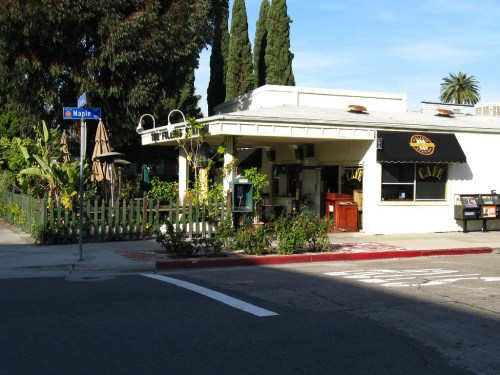 A cozy café on a street corner, surrounded by greenery and tall trees, with a sign reading "The Friendly Café."