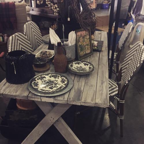 A rustic wooden table set with decorative plates, a vase, and various kitchen items in a cozy shop setting.