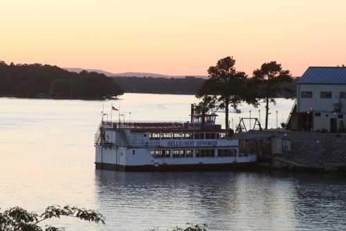 A riverboat docked at sunset, with calm waters and trees lining the shore in the background.