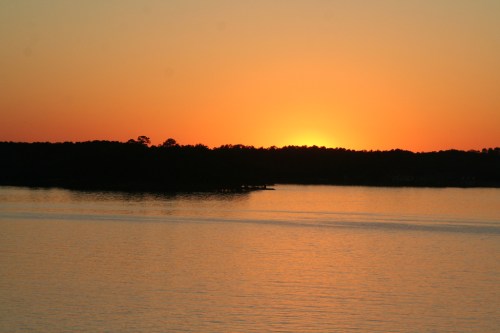 A serene sunset over a calm lake, with silhouettes of trees along the horizon and warm orange and yellow hues in the sky.