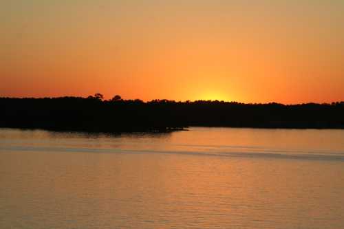 A serene sunset over a calm lake, with silhouettes of trees along the horizon and warm orange and yellow hues in the sky.