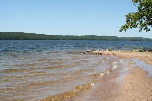 A serene beach scene with gentle waves lapping at the shore, surrounded by green hills under a clear blue sky.