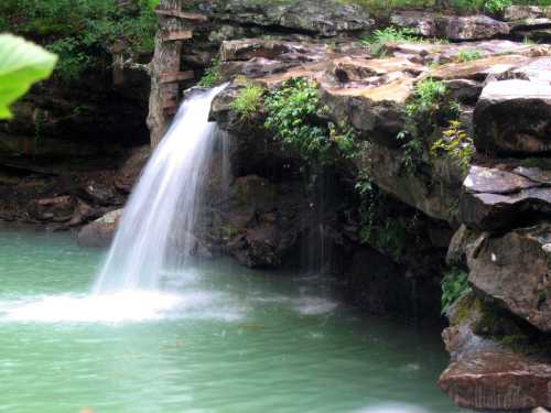 A serene waterfall cascades over rocky cliffs into a tranquil turquoise pool surrounded by lush greenery.