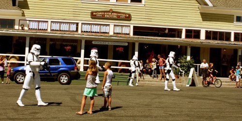 Stormtroopers walk in front of a hotel while children and families watch in a lively street scene.
