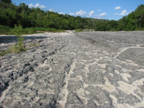 A rocky, dry riverbed with textured gray stone, surrounded by green trees and blue sky with scattered clouds.