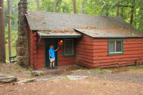 A woman in a blue shirt stands by the door of a rustic red cabin surrounded by trees and a calm lake.