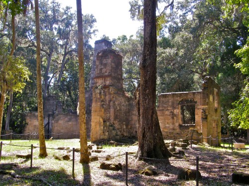 Ruins of a stone structure surrounded by trees and greenery, with a clear blue sky above.