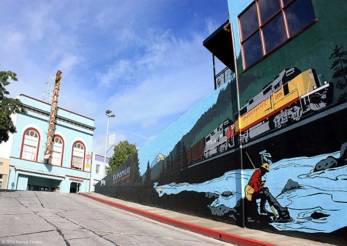 A mural depicting a train and a person fishing, with a vintage theater in the background under a blue sky.