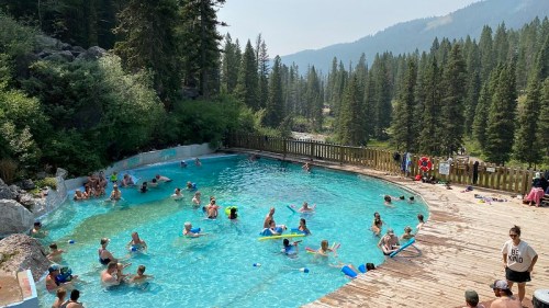 A crowded outdoor pool surrounded by trees and mountains, with people swimming and relaxing in the water.
