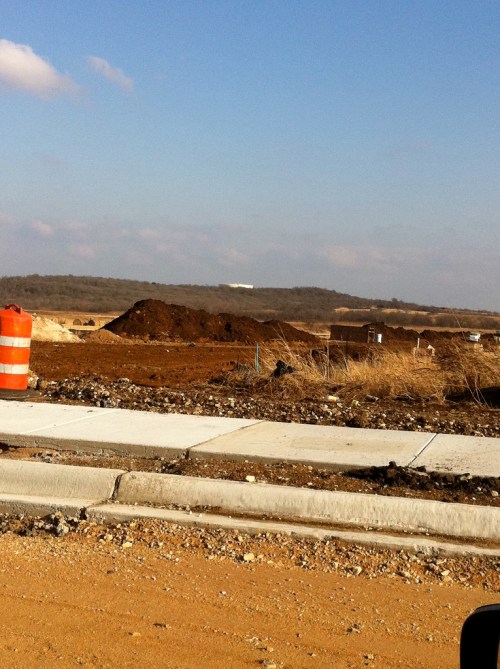 A construction site with dirt mounds, a clear blue sky, and a distant hill in the background.