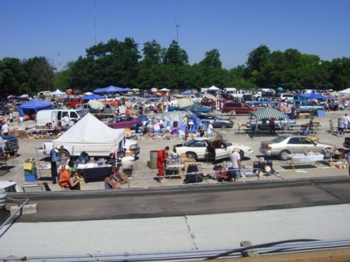 A bustling outdoor flea market with numerous tents, cars, and people browsing various items under a clear blue sky.