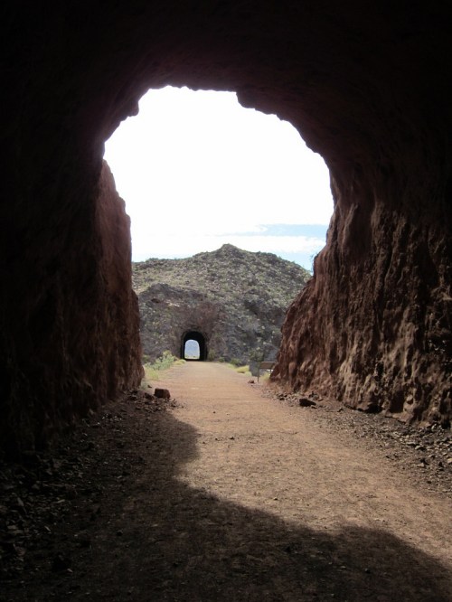 A view through a rocky tunnel, leading to another opening in the distance, with a clear sky beyond.