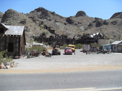 Abandoned mining town with old buildings, vintage vehicles, and mountains in the background under a clear blue sky.