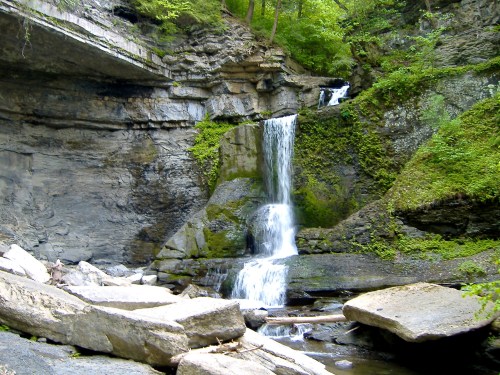 A serene waterfall cascades over moss-covered rocks, surrounded by lush greenery and rugged cliffs.