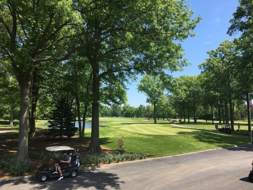 A sunny golf course with lush green trees, a pond, and a golf cart parked on a path.