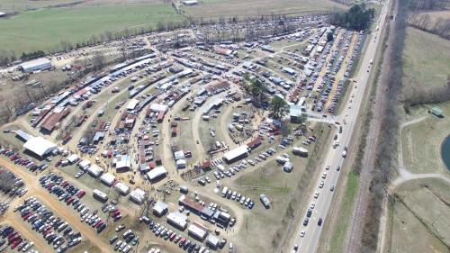 Aerial view of a large outdoor market with numerous stalls and vehicles, surrounded by fields and a road.