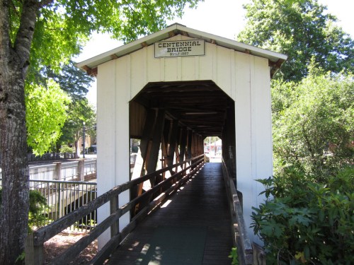 A covered wooden bridge with a sign reading "Centennial Bridge," surrounded by trees and greenery.