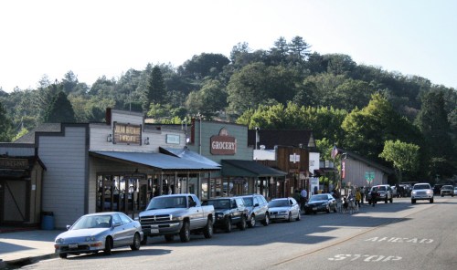 A quaint street lined with shops and parked cars, set against a backdrop of green hills and trees.